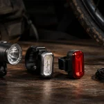 Three bike lights arranged on a wooden table beside a loaded gravel bike