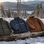 Three insulated hiking jackets laid out on a snowy log beside a daypack