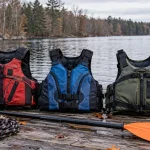 Three kayaking life jackets laid out on a wooden dock beside a paddle